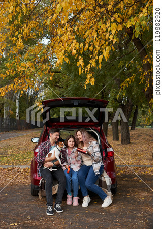 Happy parents with daughter and cute dog Jack Russel terrier sitting in car trunk on autumn day 119882920