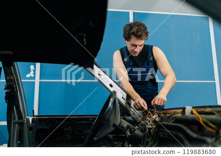 Focused mechanic in blue overalls repairing car electrical system in repair garage, troubleshooting wiring with precision. Concept of professional car repair and maintenance work. 119883680