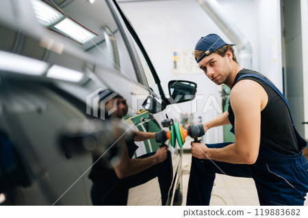 Portrait of confident mechanic male in uniform polish auto door using grinding unit in automobile repair and renew service station. Repair after accident. Concept of car repair and maintenance work. 119883682