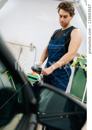 Professional muscular repairman in uniform polishing paintwork hood of car. Vertical shot of auto mechanic worker using grinding unit to polish hood at automobile repair and renew service station. 119883687