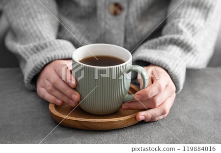 Woman in cozy sweater holding mug with espresso coffee close up on a gray table close up. 119884016
