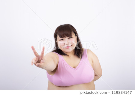 Fat young woman in a sports bra smiling and making a V sign in front of a white background Fat young woman in a sports bra smiling and making a V sign in front of a white background 119884147