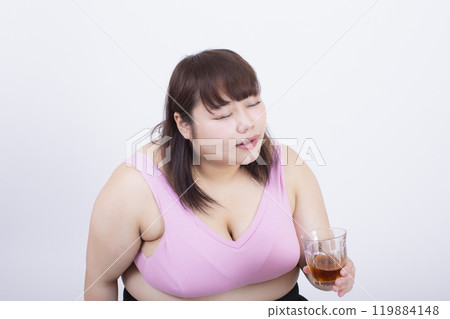 Fat young woman in a sports bra drinking a drink from a cup with relish in front of a white background Fat young woman in a sports bra drinking a drink from a cup with relish in front of a white background 119884148