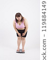 一位穿著運動胸罩的胖年輕女子站在體重計上,在白色背景前環顧四周 一位穿著運動胸罩的胖年輕女子站在體重計上,在白色背景前環顧四周 119884149