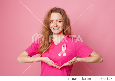 Close up of smiling young woman with pink ribbon pinned to her shirt symbolizing breast cancer awareness and importance of early detection. Healthcare concept 119884197