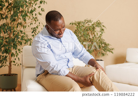 Knee pain sick body health care concept A young man has knee pain, sitting on the sofa at home, touching his leg, feeling pain in his knee. Knee pain sick body health care concept A young man has knee pain, sitting on the sofa at home, touching his leg, feeling pain in his knee. 119884222