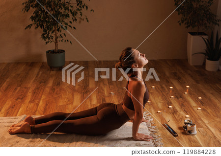 A woman doing yoga and meditation in her room doing the cobra pose, surrounded by candles and plants, creating a calming atmosphere. 119884228
