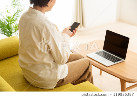 中高級女性坐在家裡客廳的沙發上，使用電腦和智慧型手機 119886397