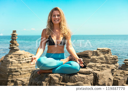 Yoga woman sitting in lotus position cliff rock beach sea blue sky 119887770
