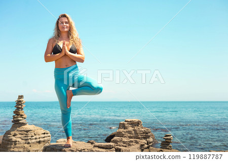 Yoga woman standing in vrikshasana tree pose hand namaste on cliff at sea sky horizon 119887787