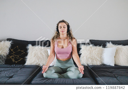 Smiling yoga woman in headphones sitting in lotus position on couch enjoy meditation Smiling yoga woman in headphones sitting in lotus position on couch enjoy meditation 119887842