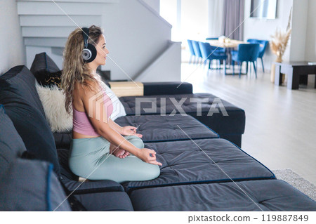 Smiling yoga woman in headphones sitting in lotus position on couch enjoy meditation 119887849