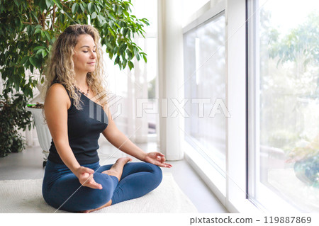 Woman yoga meditation sitting in lotus position near panorama window with houseplant at home 119887869
