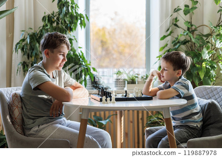 Teenage boy and his little brother play chess at a round white table on a chessboard. C Teenage boy and his little brother play chess at a round white table on a chessboard. C 119887978