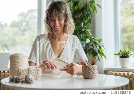 A young girl in a white dress sits at a table and knits a basket from beige yarn. 119888248