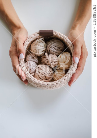 Female hands holding a crocheted basket with skeins of cotton yarn in natural tones and colors. 119888263