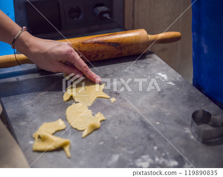 Female pastry chef skillfully uses star-shaped cookie cutter to create delightful cookies from cookie dough on gray kitchen counter. Nearby, wooden rolling pin rests, creations and delicious treats. Female pastry chef skillfully uses star-shaped cookie cutter to create delightful cookies from cookie dough on gray kitchen counter. Nearby, wooden rolling pin rests, creations and delicious treats. 119890835
