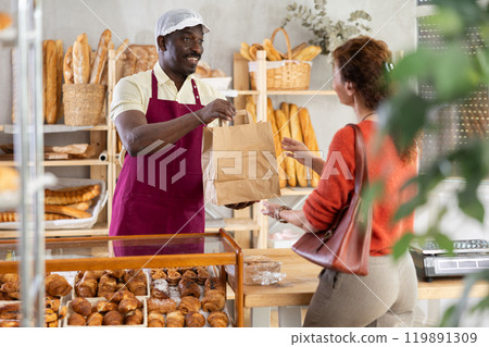 Helpful baker salesman hands paper bag with hot delicious baguettes to female customer 119891309