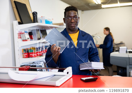 Middle-aged African American man in blue robe uniform smiling at the camera and holding new planners in printer house Middle-aged African American man in blue robe uniform smiling at the camera and holding new planners in printer house 119891870