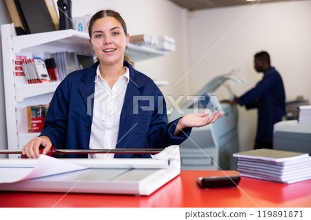 Joyful young woman in a blue uniform smiling at the camera and using paper cutter in printer house 119891871