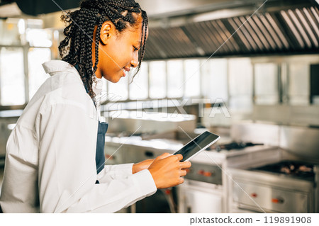 Female chef in world famous restaurant's modern kitchen cooks using tablet. Gourmet organic dishes prepared technology assists. Professional smiling reading communicating. Female chef in world famous restaurant's modern kitchen cooks using tablet. Gourmet organic dishes prepared technology assists. Professional smiling reading communicating. 119891908