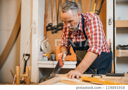 A carpenter in a red plaid shirt and blue overalls saws a piece of wood with a handsaw on a workbench, concentrating on precise cuts in the woodworking project, National Carpenters Day 119891916