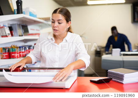 Concentrated young woman in white shirt using paper cutter on the table with planners and calculator in the printer house Concentrated young woman in white shirt using paper cutter on the table with planners and calculator in the printer house 119891960