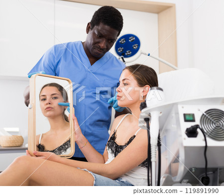 Female client looking at mirror while beautician examining her face 119891990