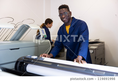 Positive middle-aged African American man in uniform using plotter during work in the printing office 119891998
