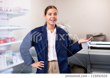 Cheerful young woman in blue robe uniform sticking out her left hand to show the workflow in the printer house Cheerful young woman in blue robe uniform sticking out her left hand to show the workflow in the printer house 119892026