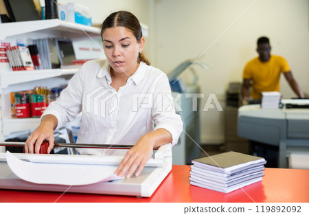 Focused young female typographer trimming the printed work on the cutter in the typography Focused young female typographer trimming the printed work on the cutter in the typography 119892092