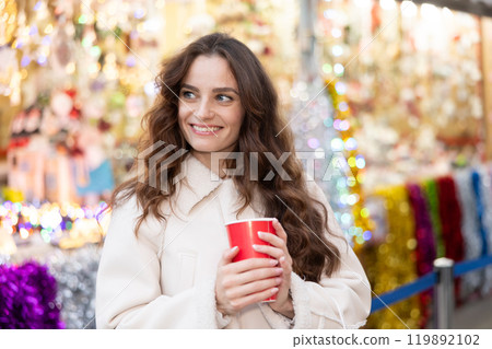 Smiling woman walking through Christmas street market with warm beverage in paper cup Smiling woman walking through Christmas street market with warm beverage in paper cup 119892102