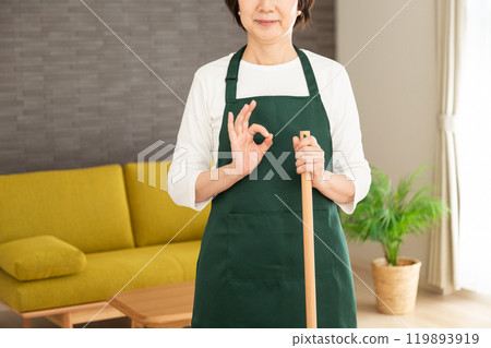 A senior female housewife/housekeeper wearing a green apron smiling and making a circle sign in the living room at home 119893919