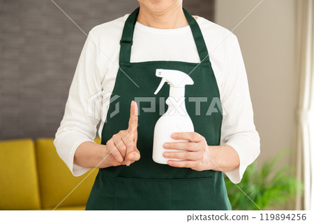 A senior female housewife/housekeeper wearing a green apron pointing up in the living room at home A senior female housewife/housekeeper wearing a green apron pointing up in the living room at home 119894256