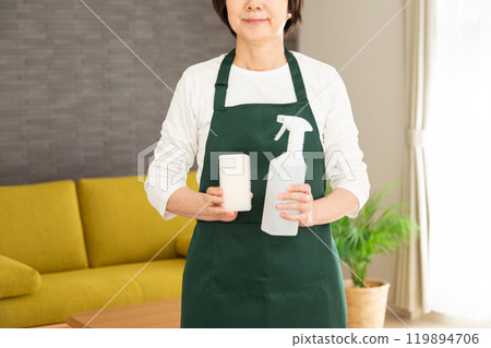 A senior female housewife/housekeeper wearing a green apron smiling with detergent and a sponge in her hand in the living room at home A senior female housewife/housekeeper wearing a green apron smiling with detergent and a sponge in her hand in the living room at home 119894706