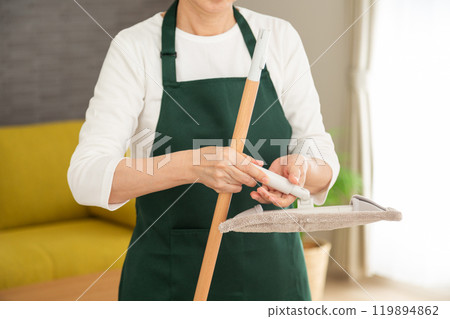 A senior female housewife/housekeeper wearing a green apron holding a mop in the living room at home 119894862