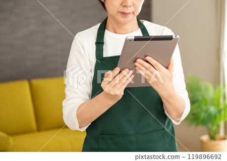 一位身穿綠色圍裙的高級女家庭主婦/管家看著自家客廳裡的平板電腦 一位身穿綠色圍裙的高級女家庭主婦/管家看著自家客廳裡的平板電腦 119896692