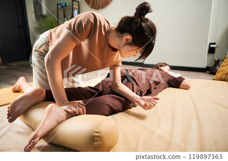 Masseuse making thai yoga massage. Woman therapist doing traditional massage treatment for female patient using elbow. Client lying on mat, meditating, enjoying hand massage in calm atmosphere. 119897363