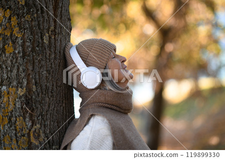 Portrait of a beautiful senior woman with headphones in autumn park, listening music with her eyes closed. Portrait of a beautiful senior woman with headphones in autumn park, listening music with her eyes closed. 119899330