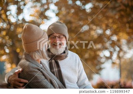 Portrait of beautiful senior couple sitting on bench in autumn park. Elderly husband and wife looking at each other lovingly. 119899347