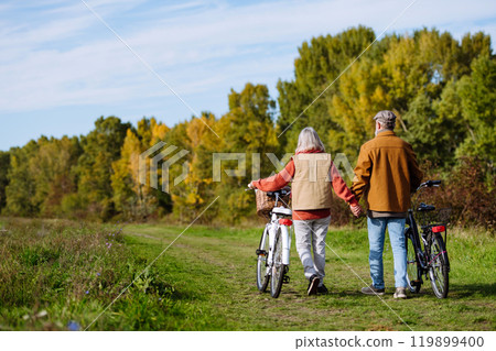 Rear view of elderly couple on a walk in the forest, pushing their bikes side by side. Seniors in love on stroll through autumn nature, enjoying a peaceful moment together. Rear view of elderly couple on a walk in the forest, pushing their bikes side by side. Seniors in love on stroll through autumn nature, enjoying a peaceful moment together. 119899400