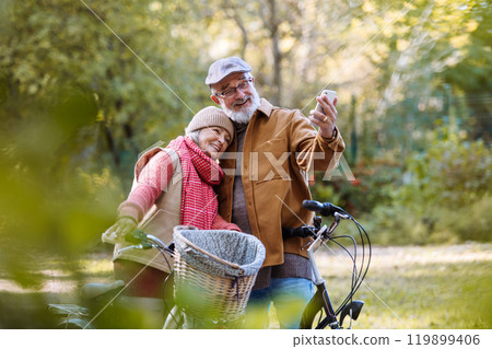 Lovely elderly couple on standing in the middle of autumn nature with their bikes, taking selfie with smartphone. 119899406