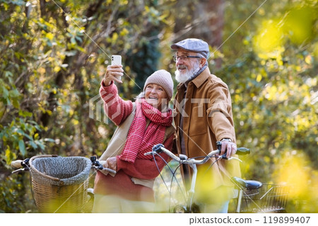 Lovely elderly couple on standing in the middle of autumn nature with their bikes, taking selfie with smartphone. 119899407