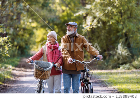 Lovely elderly couple on a walk in the forest, pushing their bikes side by side. Seniors in love on stroll through autumn nature, enjoying a peaceful moment together. 119899408