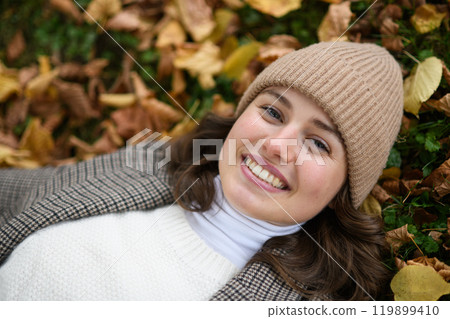 Portrait of a beautiful woman lying in fallen autumn leaves on the lawn. She smiles and looking at camera. 119899410