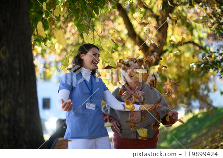 Portrait of nurse and elderly woman on walk in the park during warm autumn day, playing with fallen colorful leaves. 119899424