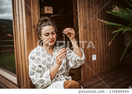 Beautiful woman doing her skin care routine outdoors, wearing bathrobe. Woman using facial skin serum with dropper. Beautiful woman doing her skin care routine outdoors, wearing bathrobe. Woman using facial skin serum with dropper. 119899444