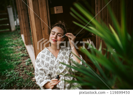Beautiful woman doing her skin care routine outdoors, wearing bathrobe. Woman using facial skin serum with dropper. Beautiful woman doing her skin care routine outdoors, wearing bathrobe. Woman using facial skin serum with dropper. 119899450