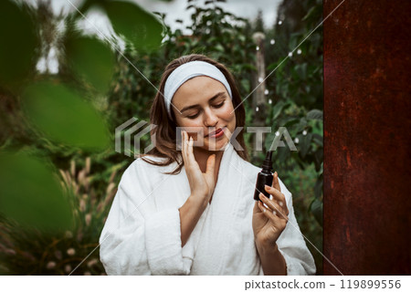 Beautiful woman doing her skin care routine outdoors, wearing bathrobe. Woman using facial skin serum with dropper. Beautiful woman doing her skin care routine outdoors, wearing bathrobe. Woman using facial skin serum with dropper. 119899556