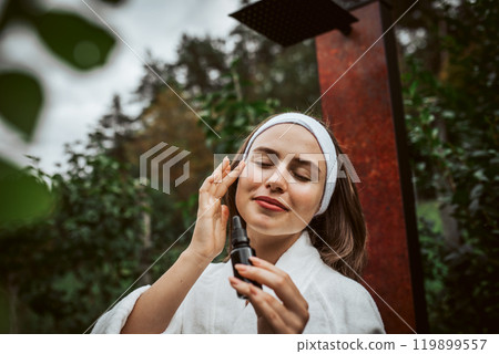 Beautiful woman doing her skin care routine outdoors, wearing bathrobe. Woman using facial skin serum with dropper. Beautiful woman doing her skin care routine outdoors, wearing bathrobe. Woman using facial skin serum with dropper. 119899557